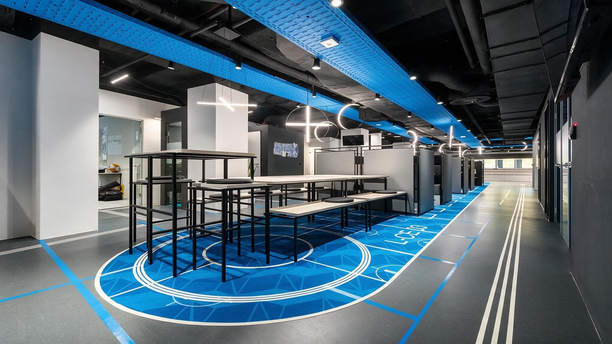 Weitwinkelblick über die blaue Community-Fläche im MerkurOffice: Hochtische auf dem Kreis-Teppich, Laufbahn-Markierungen und blau beleuchtete Decke